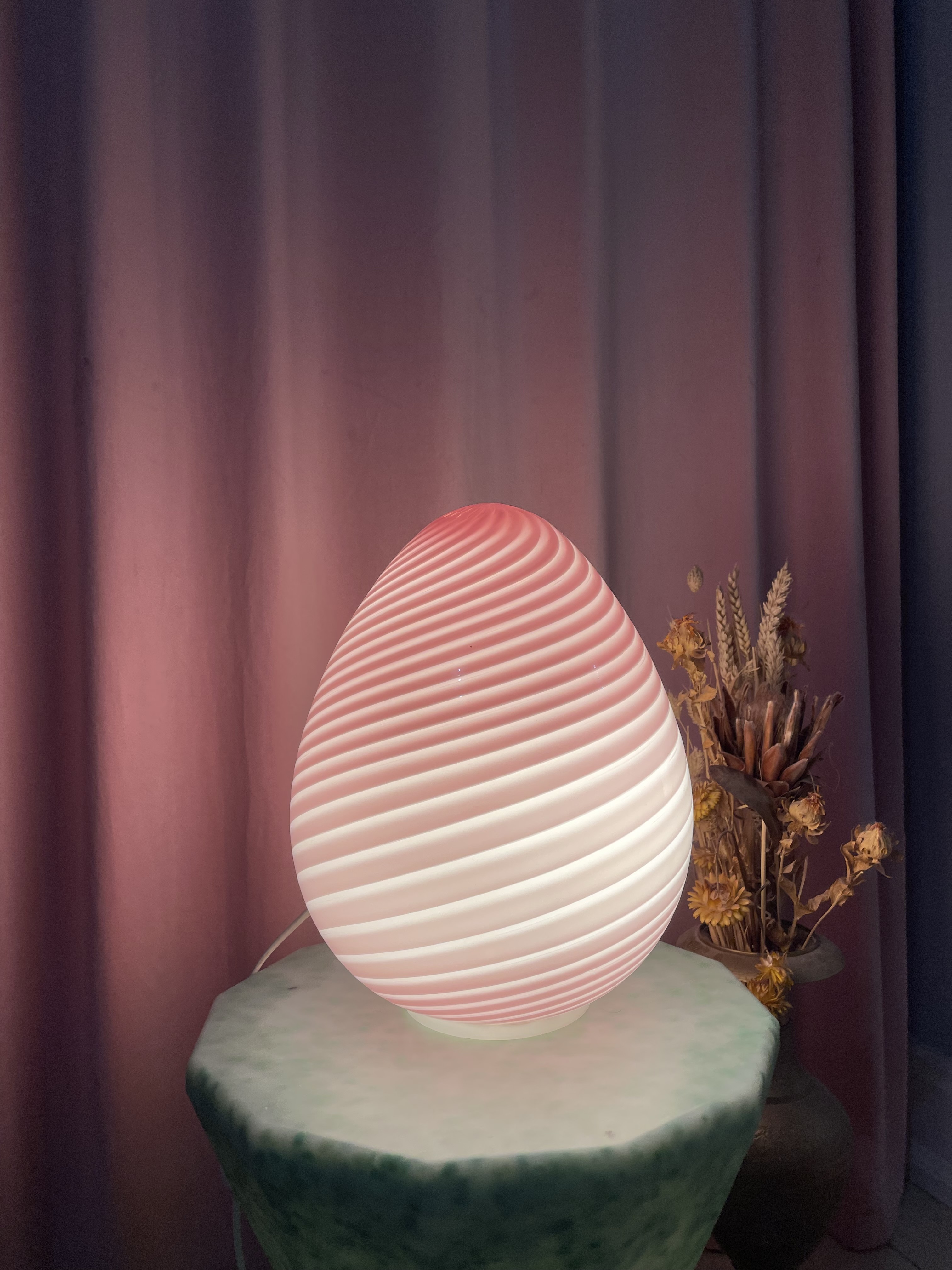 Tændt lyserød Murano bordlampe med swirl-mønster på et sidebord foran et gardin
