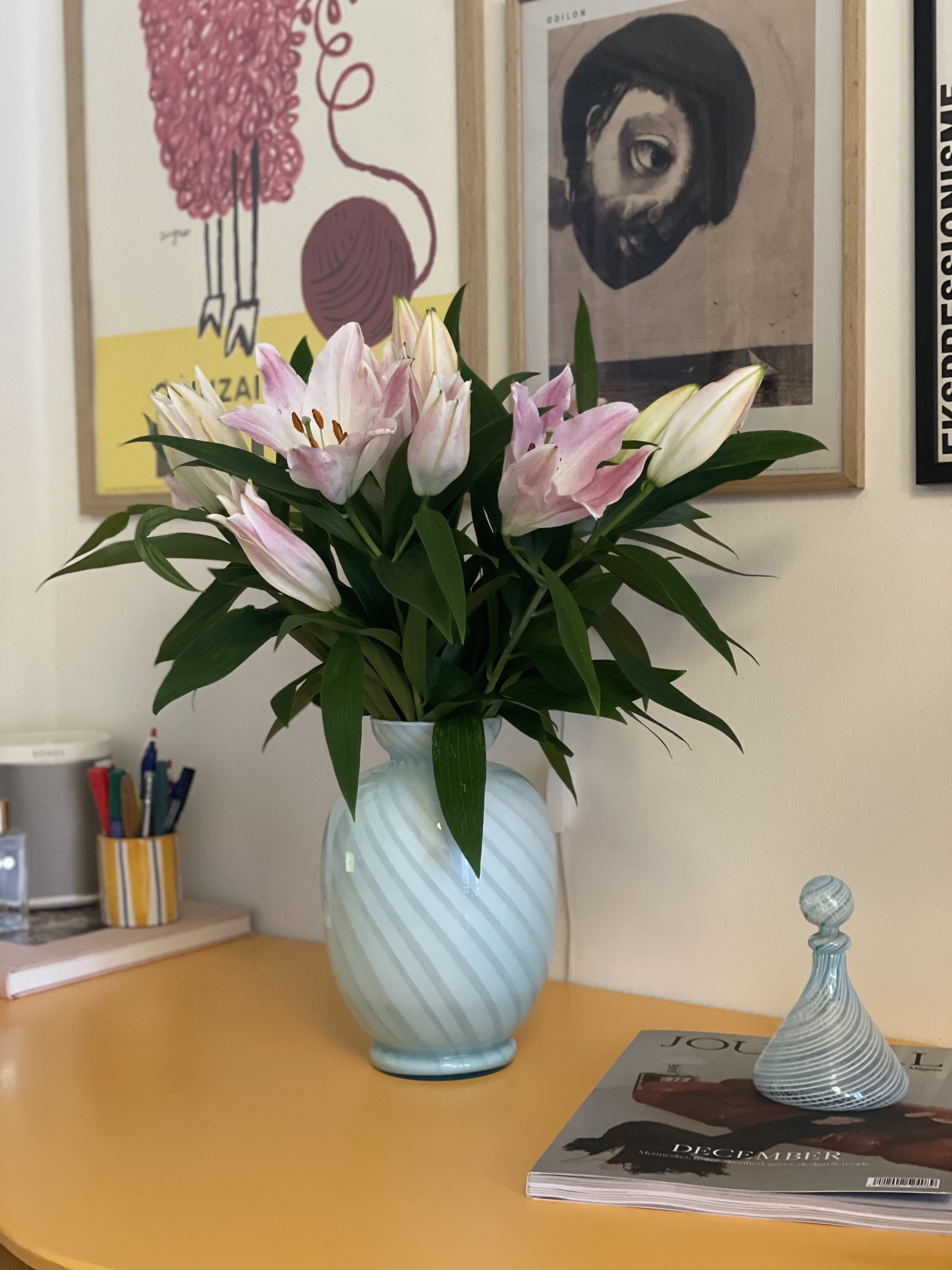 Lyseblå Murano vase med swirl-mønster og lyserøde liljer på et gult bord